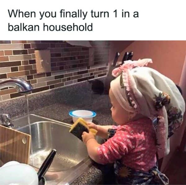Balkan meme showing baby in headscarf standing on stool washing dishes at kitchen sink.
