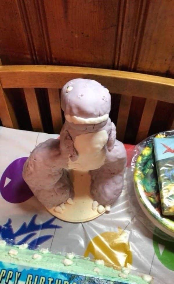 Purple frosted dinosaur cake sagging on a table beside party plates and a birthday banner