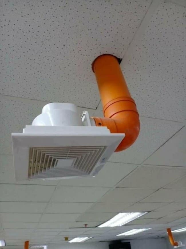 Orange duct pipe bending out of a ceiling tile to feed a vent upside down, pure construction fails.