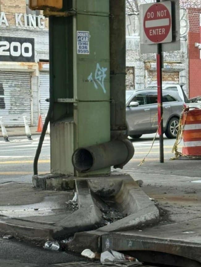 Pipe on a street pole draining straight into a crumbling curb beside a do not enter sign.