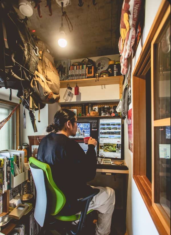 Man working at a narrow desk in a tiny room crammed with camping gear and shelves
