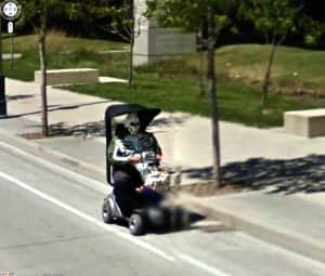 Person in a skeleton mask riding a mobility scooter down a sidewalk on street view.