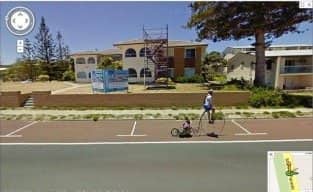 Person pushing a tiny wheeled cart down a seaside street past a scaffolded house.