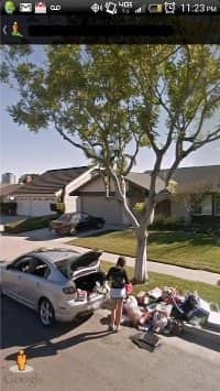 Woman unloading a car trunk onto a curbside pile of belongings on a suburban street.