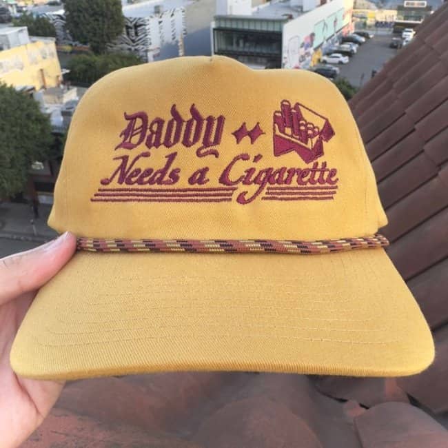 Vintage style yellow trucker hat with embroidered red text saying Daddy Needs a Cigarette next to a cigarette pack logo.