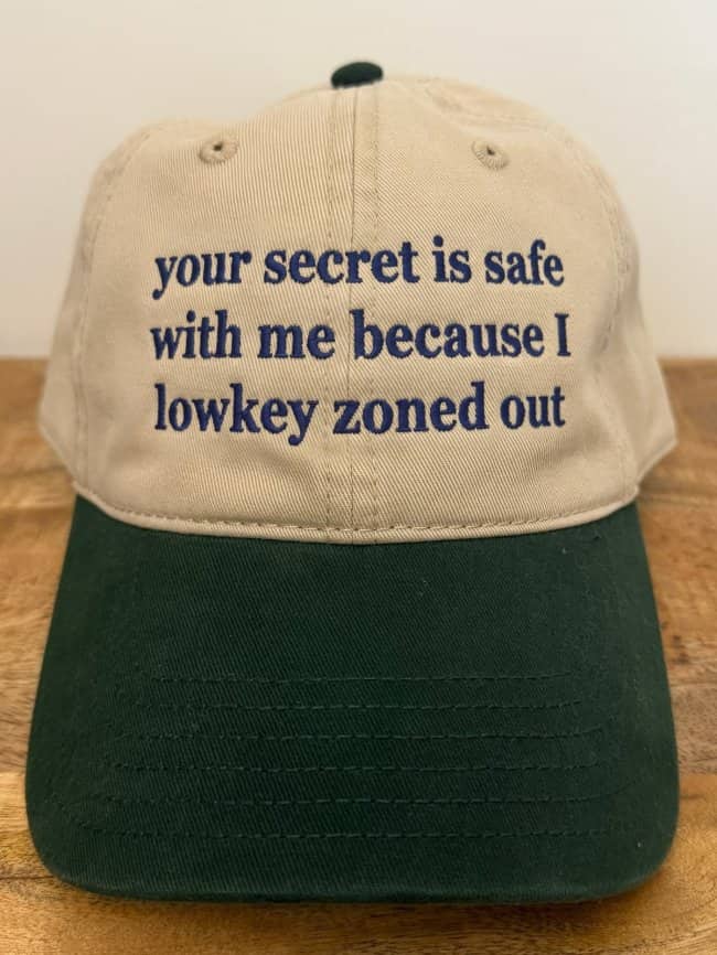 Two-tone tan and green baseball cap embroidered with text reading your secret is safe with me because I lowkey zoned out.