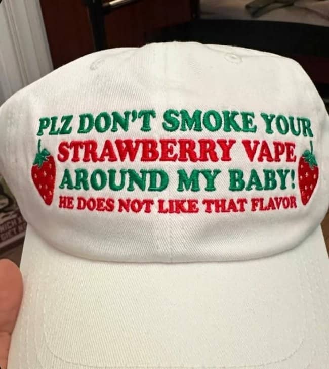 White baseball cap with green and red embroidered text asking people not to smoke strawberry vape around a baby.