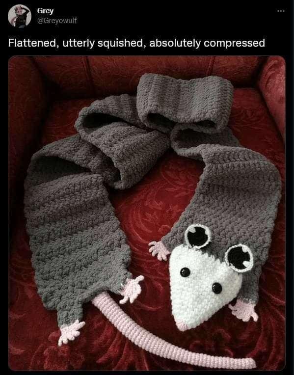 Funny memes crocheted grey opossum scarf laid flat on a red armchair cushion.
