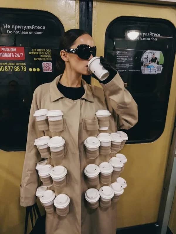 Stylish woman wearing trench coat covered in takeaway coffee cups while riding the subway.