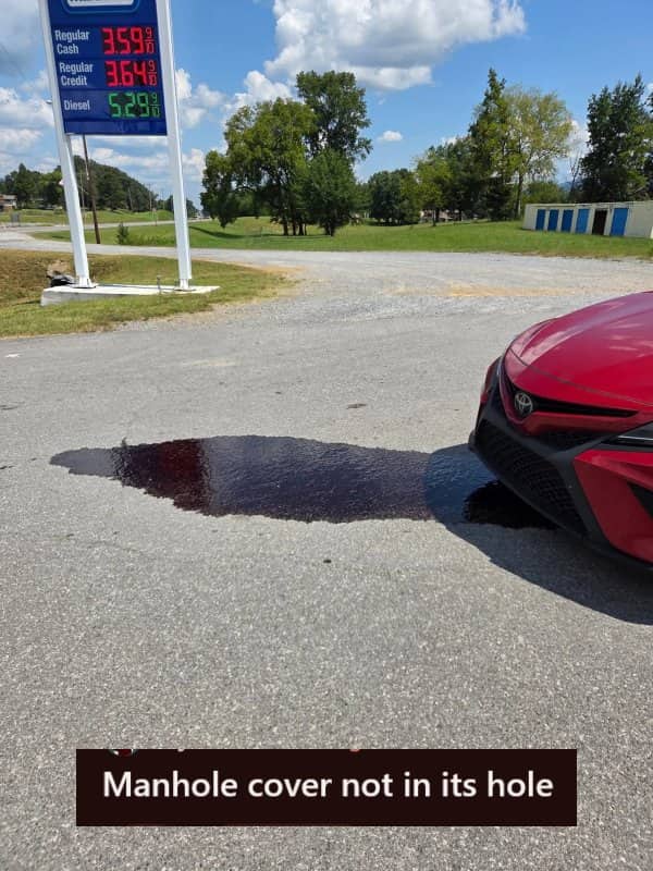 Red car leaking oil onto pavement after hitting an open manhole during a bad day.