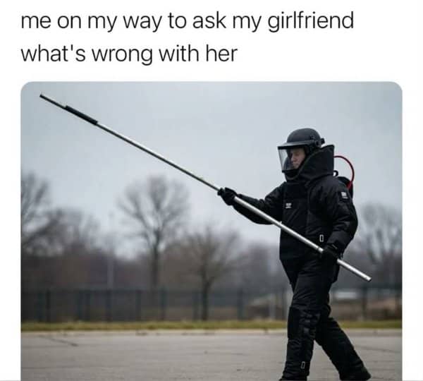Person in a full bomb disposal suit carries a long pole across an empty lot.