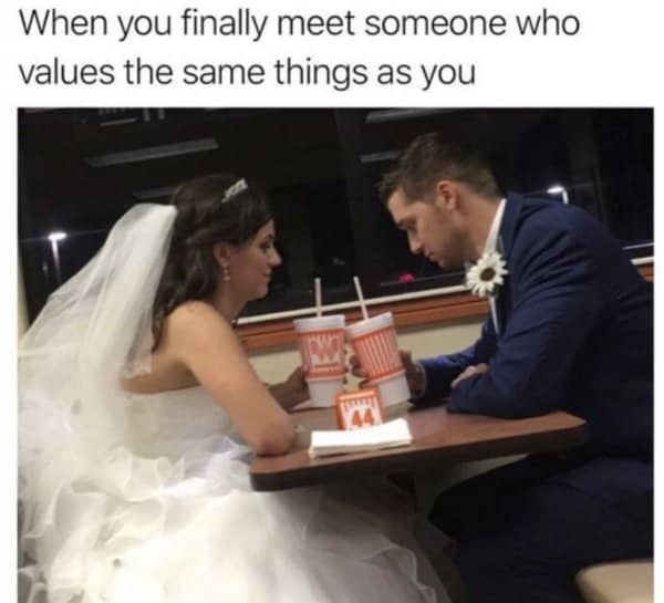 Bride and groom in full wedding attire share fast food drinks at a burger place.