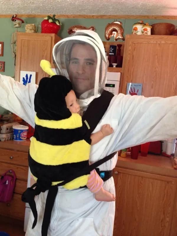 A wholesome meme dump photo of a father wearing a full white beekeeper suit holding his infant in a fuzzy bumblebee costume.