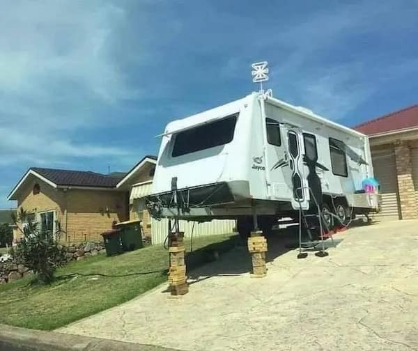 Travel trailer parked on a steep driveway dangerously propped up on tall stacks of wooden blocks in a meme dump.