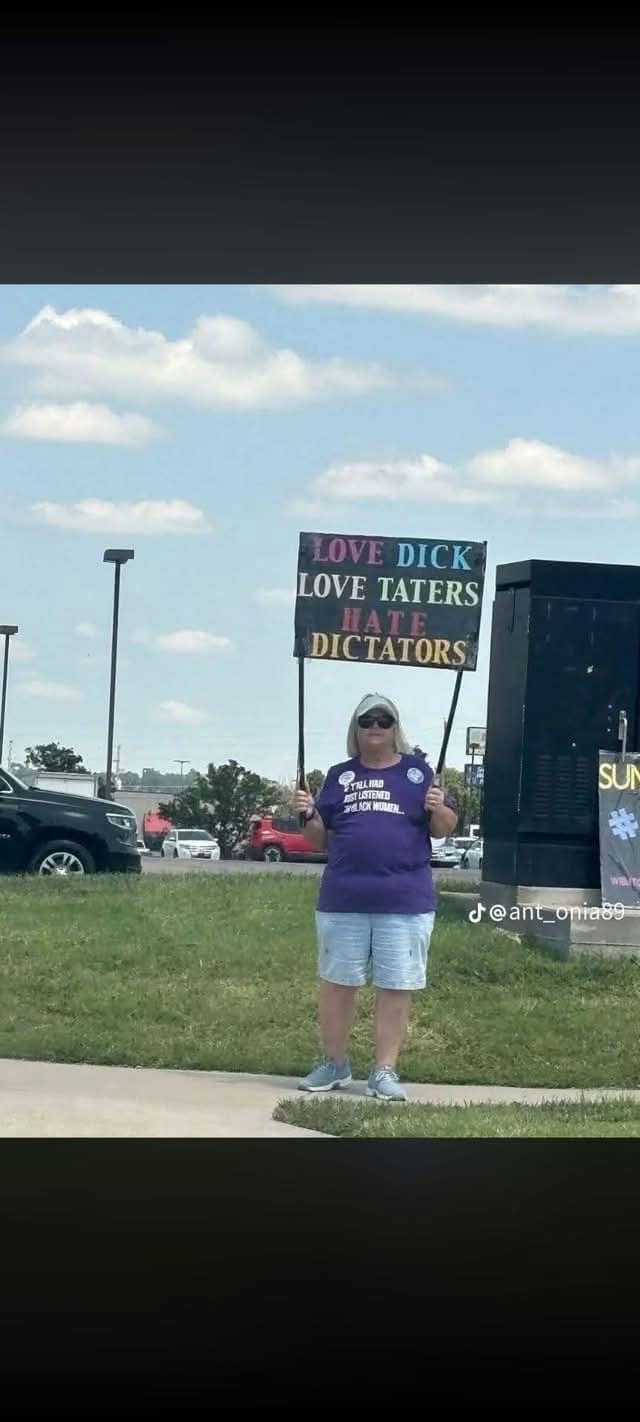 Woman protesting outdoors holding up a hand-painted poster that reads 'LOVE DICK LOVE TATERS HATE DICTATORS' in a funny meme.