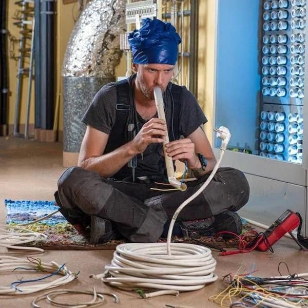 Electrician wearing a blue plastic hat playing a plastic conduit like a flute to snake-charm an electrical cable in a meme dump.