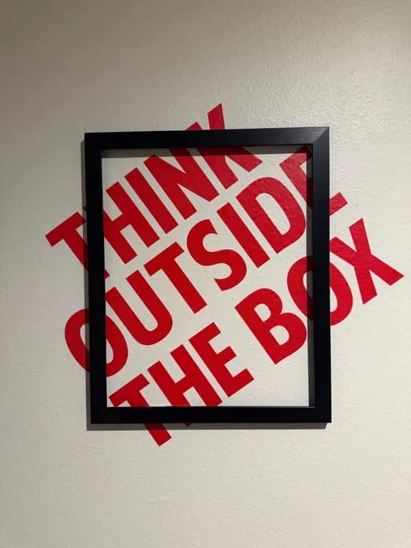 Modern art wall installation featuring a black picture frame mounted crookedly over red wall lettering reading 'THINK OUTSIDE THE BOX', shared in a funny meme.