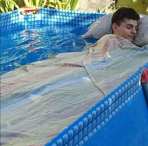 Guy sleeping peacefully tucked under a blanket directly inside a full backyard swimming pool in a meme dump.