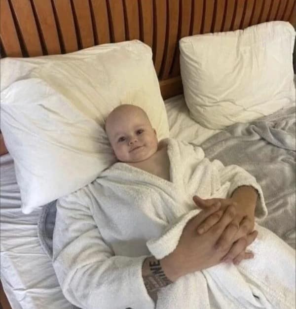 Face-swapped edit of a baby's head onto a tattooed man resting in a white bathrobe in a meme dump.