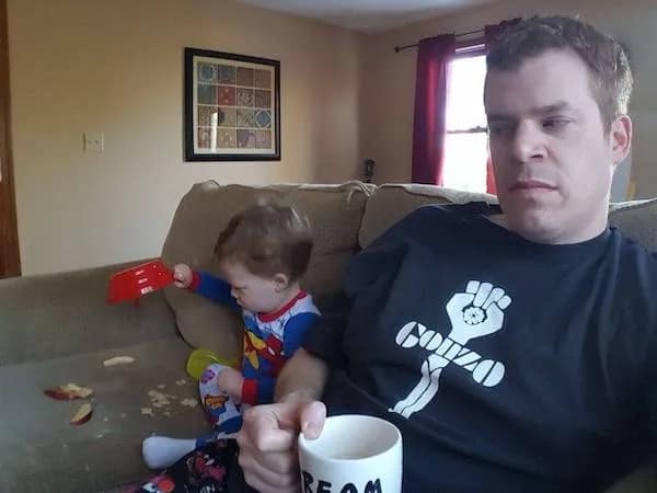 A tired father sipping coffee while his toddler dumps a red bowl of cereal onto the couch in a funny meme.