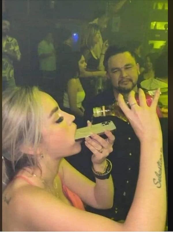 Nightclub attendee taking a tequila shot poured down the back of an iPhone held in her mouth in a funny memes capture.