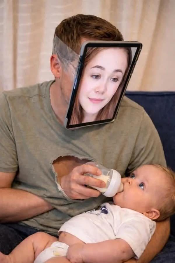 Man with a tablet of a woman's face strapped on, bottle feeding a baby.
