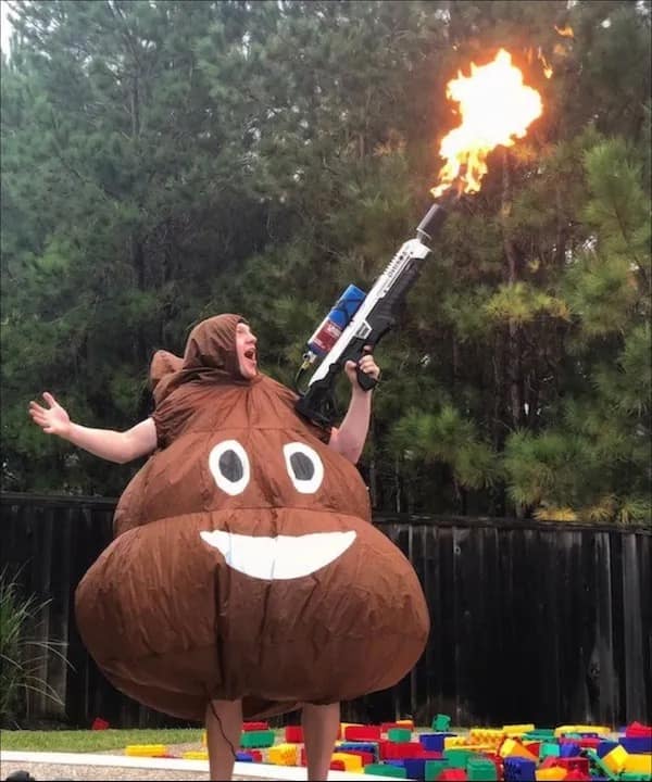 Person wearing an inflatable poop emoji costume firing a real flamethrower into the air over outdoor Lego bricks in a meme dump.