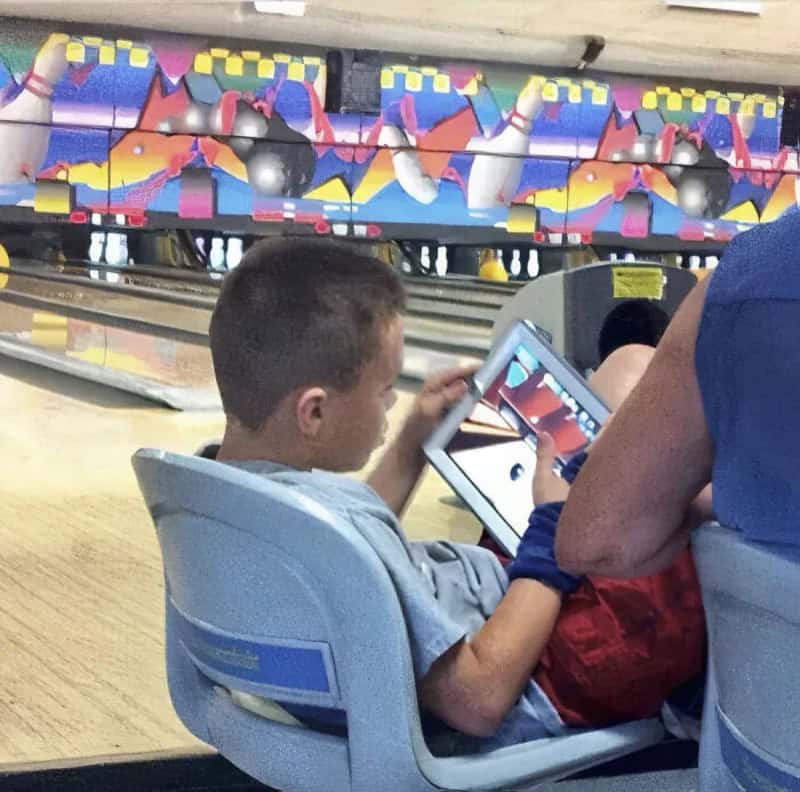 Young boy sitting in a chair at a real bowling alley lane playing a digital bowling game on his iPad in a meme dump.