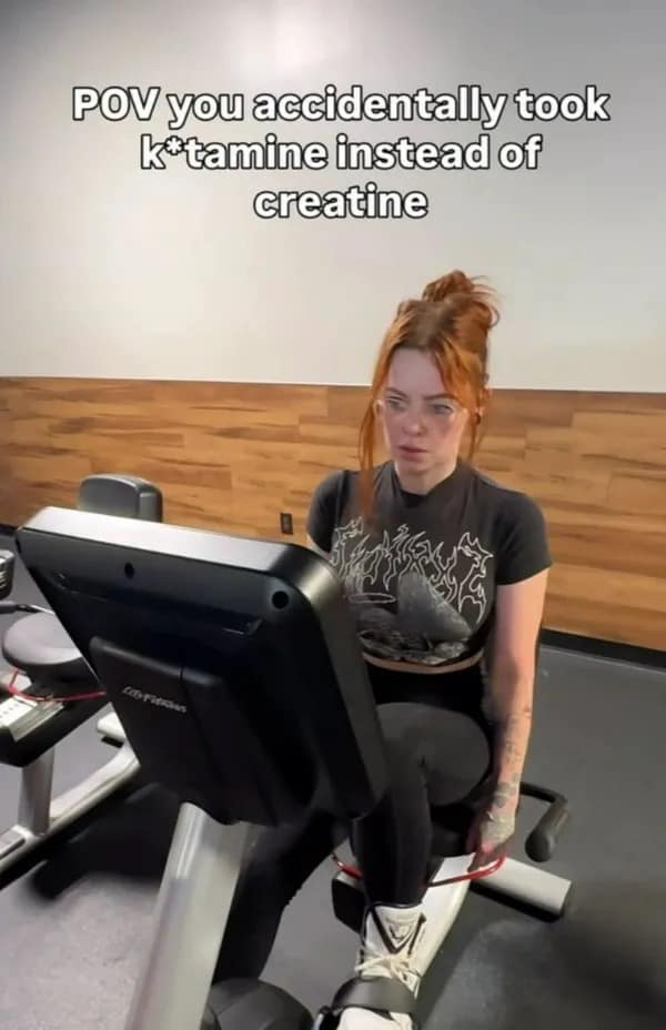Red-haired woman looking wide-eyed and thoroughly disoriented on a gym stationary bike, captioned about confusing supplements before a workout.