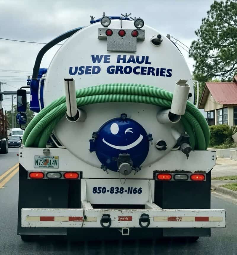 Commercial septic tank truck with a winking smiley face painted on the back and the slogan 'WE HAUL USED GROCERIES' in a meme dump.