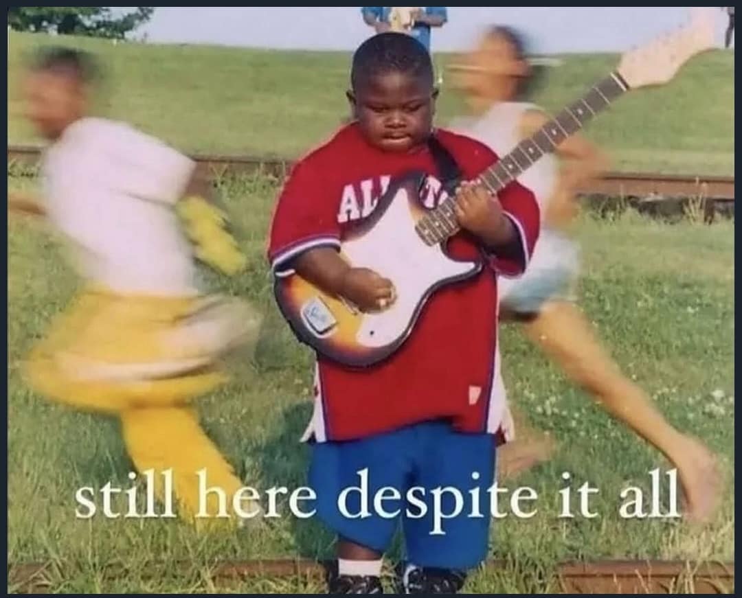 Motion-blurred photo of a young boy stoically playing electric guitar amidst chaos captioned 'still here despite it all' in a meme dump.