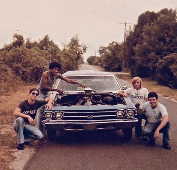 Four young men posing around vintage Chevy SS muscle car with hood open on quiet country road.