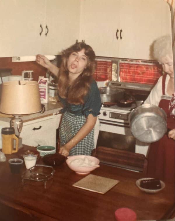 Retro photo of girl playfully sticking out tongue in kitchen while grandmother cooks dinner.