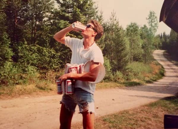 Vintage photo of man in short denim cutoffs drinking beer while carrying packs on dirt road.