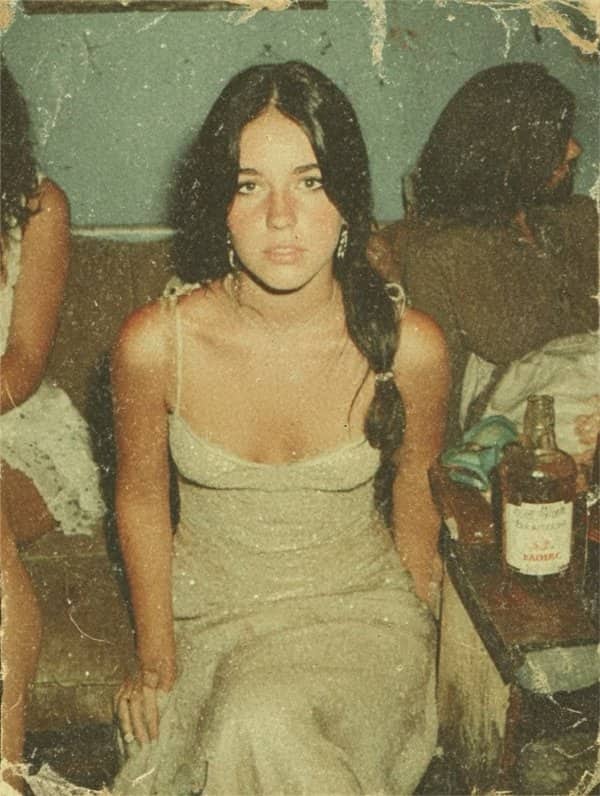 Vintage 1970s photo of young woman in summer dress sitting on couch near liquor bottle.