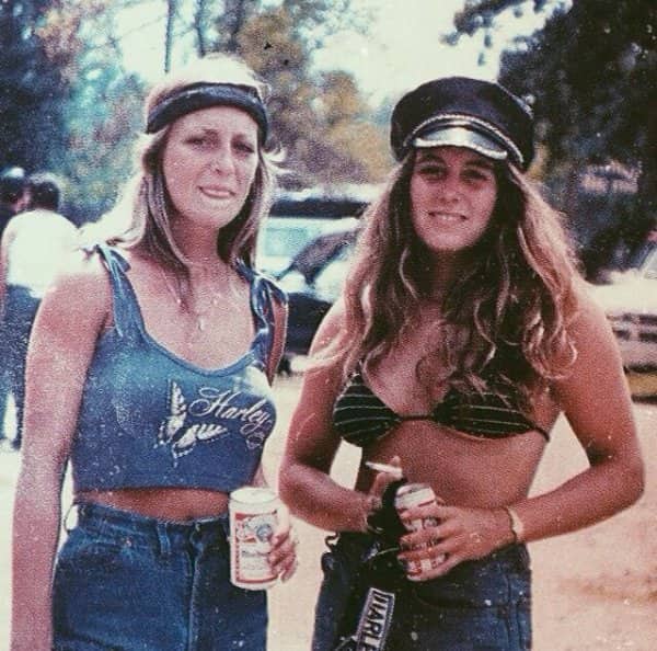 Retro photo of two women wearing Harley Davidson top and cap holding beers outdoors.