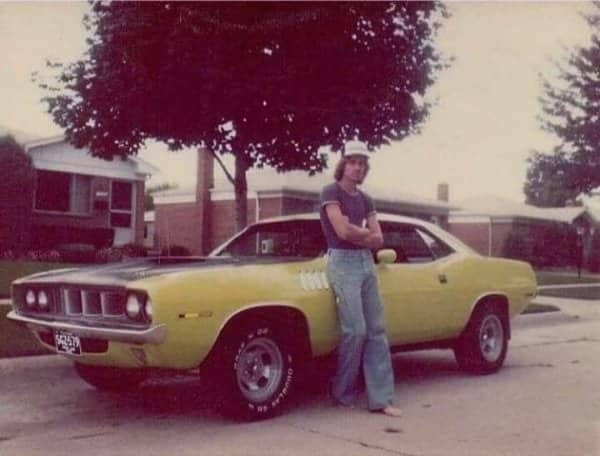 Man with long hair leaning casually against classic yellow Plymouth muscle car on suburban street.