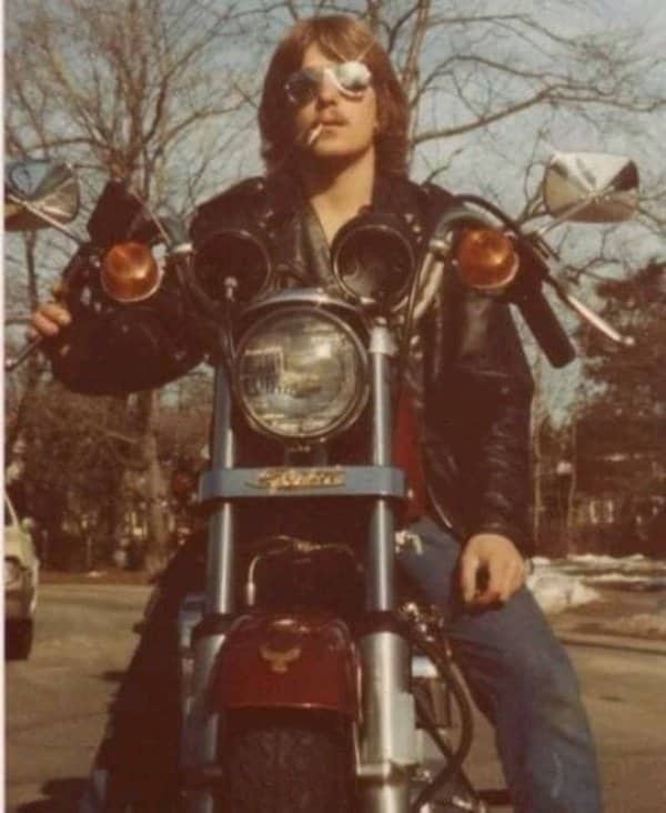 Biker in leather jacket and aviator sunglasses sitting on vintage motorcycle holding cigarette.