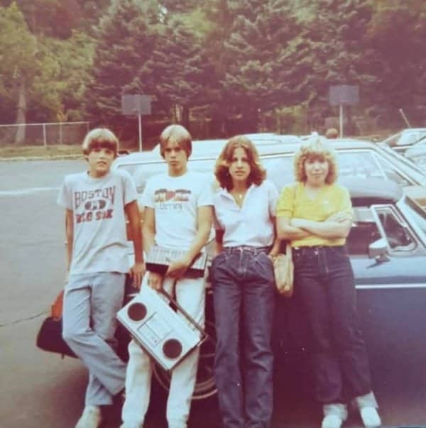 Group of teenagers posing outside leaning against car while holding large vintage boombox cassette player.