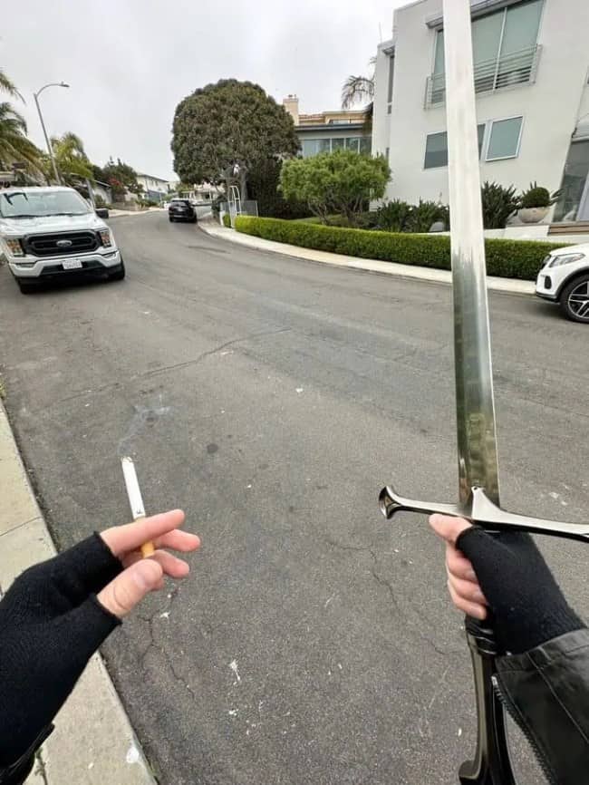 Gloved hands holding a cigarette and a long sword on a quiet suburban street.