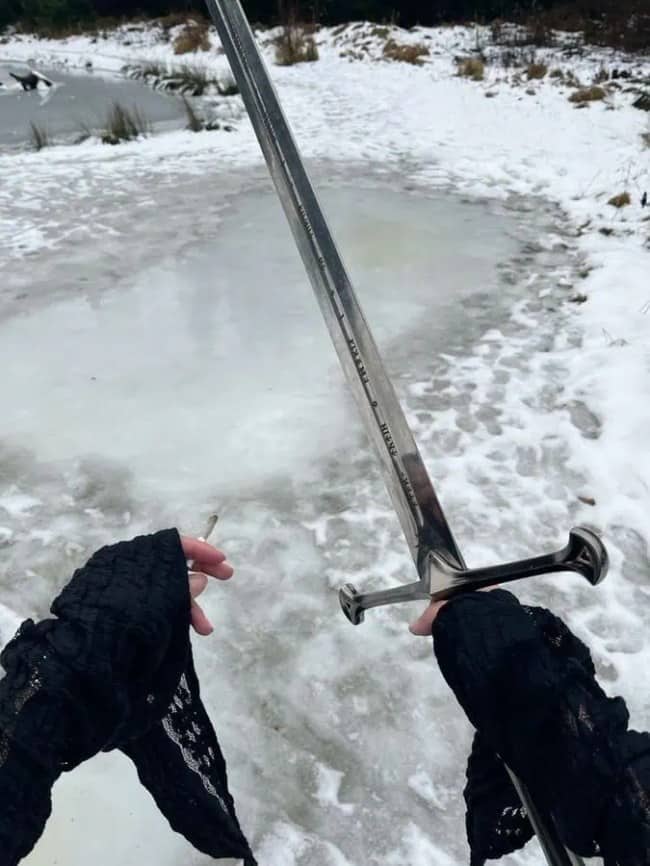 Gloved hands holding a cigarette and a sword beside a frozen pond in the snow.