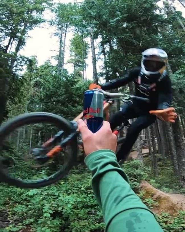 First person hand offering a Red Bull can to a mountain biker flying past in the woods.