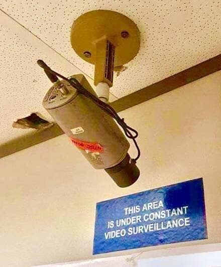 Old-school CCTV security camera mounted on a ceiling above a blue surveillance warning sign in a punny meme.