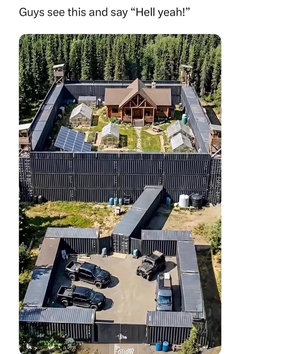 Aerial view of a log cabin compound walled in by stacked black shipping containers in a forest