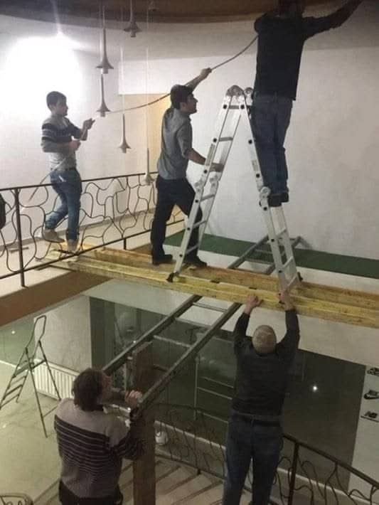 Workers balancing a ladder on a wooden plank spanning an open stairwell while others hold it