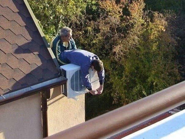 Two men leaning over a roof edge to work on an air conditioner with no harness