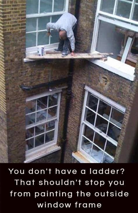 Man crouched on a plank wedged between two upper windows painting an exterior window frame