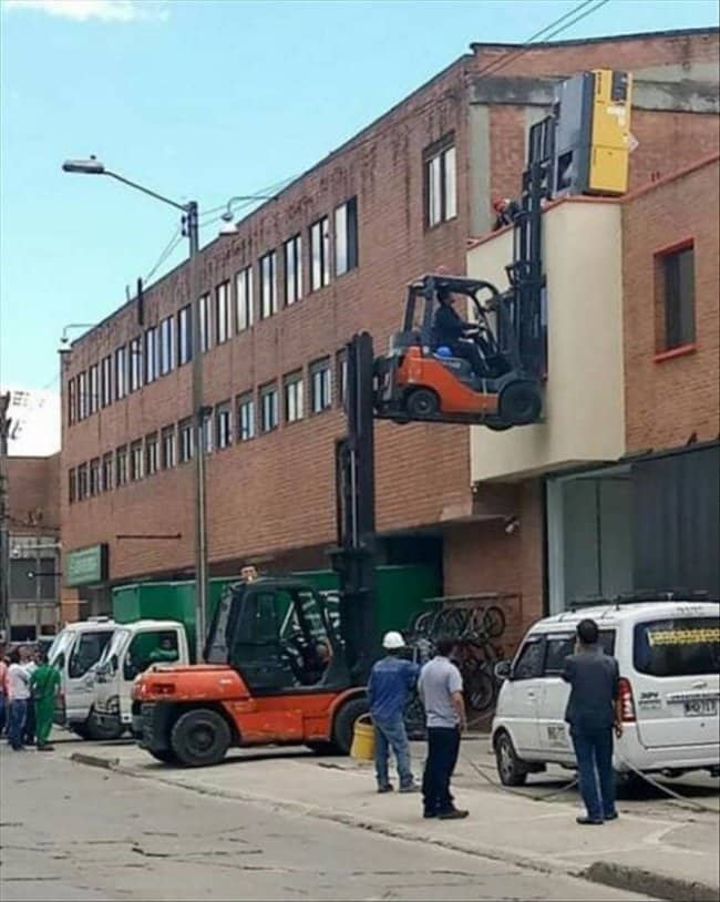 Forklift lifting a second forklift up to a second story window while onlookers watch