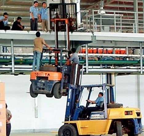 Warehouse forklift lifting another forklift with two men standing on it near a mezzanine