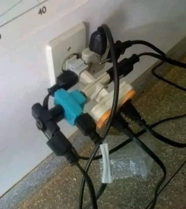 Wall outlet overloaded with stacked adapters, splitters and tangled power cables hanging down